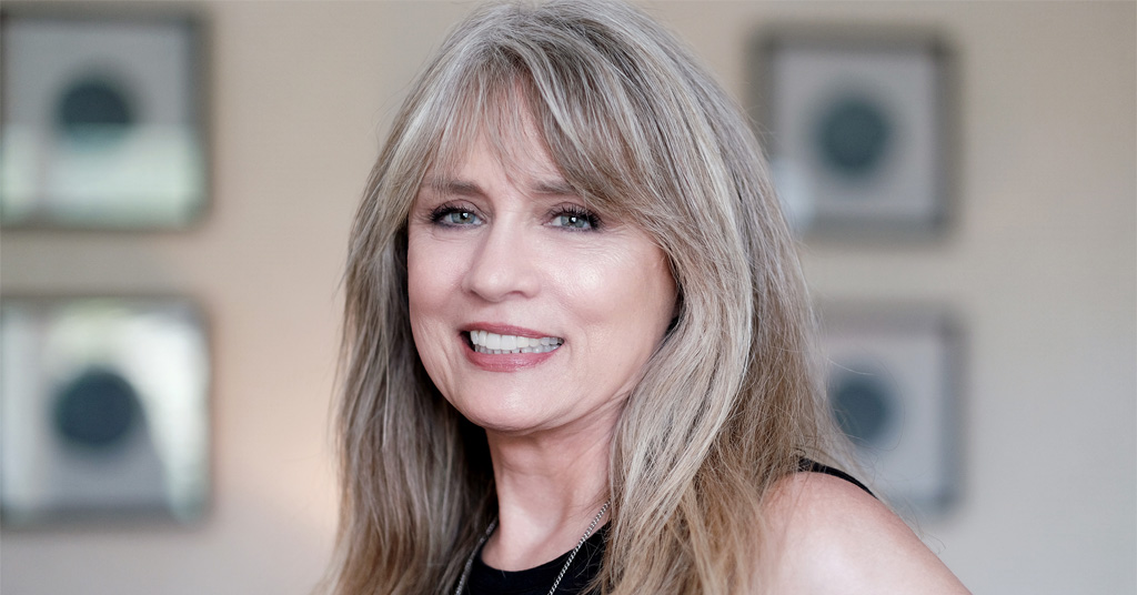 Smiling middle-aged woman with long, light brown hair standing indoors.