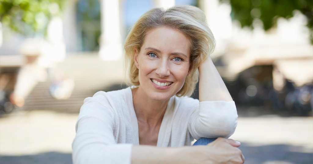 Blonde woman smiling outdoors while resting her head on her hand.