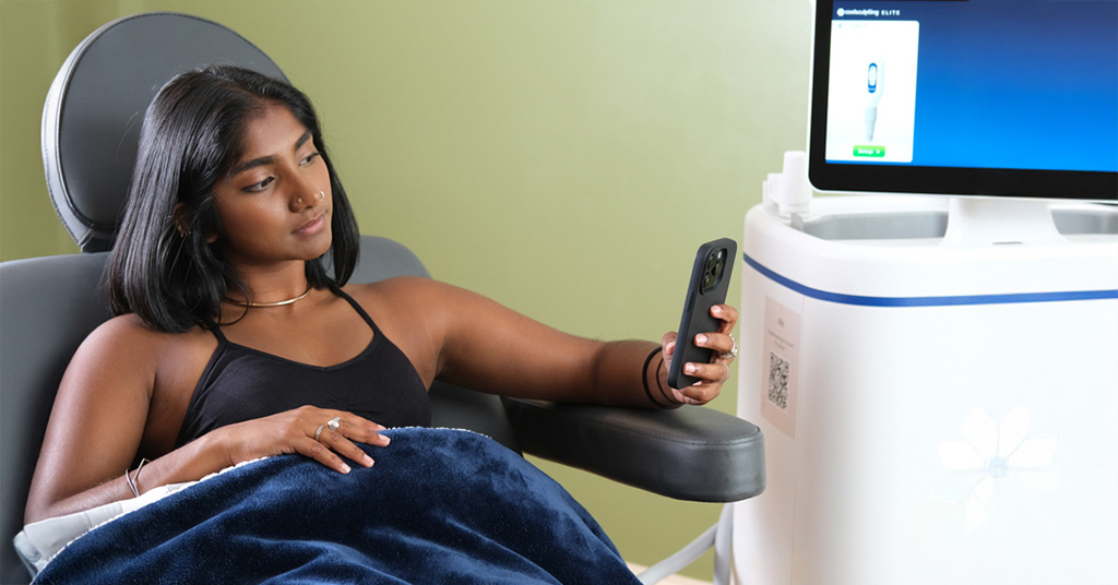 Woman reclining in a treatment chair under a blanket while looking at her phone beside a medical device.