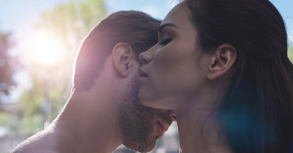 Close-up of a woman gently kissing a man’s neck in soft sunlight.