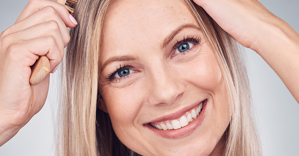 Smiling woman brushing her blonde hair while looking at the camera.