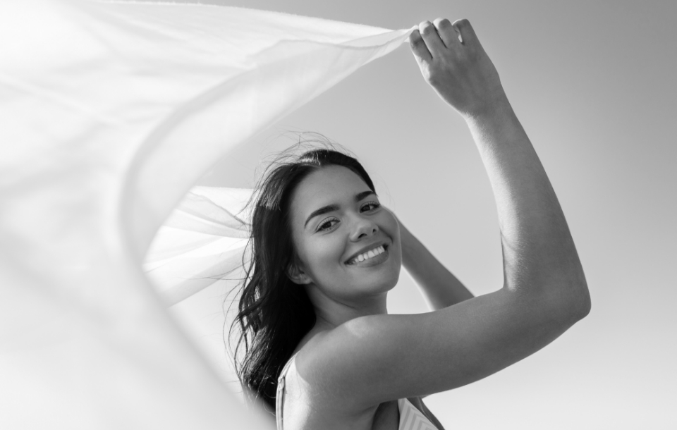 Woman smiling and holding a flowing white fabric in the breeze on a sunny day.