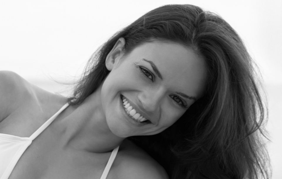 Woman smiling brightly while leaning slightly toward the camera at the beach.