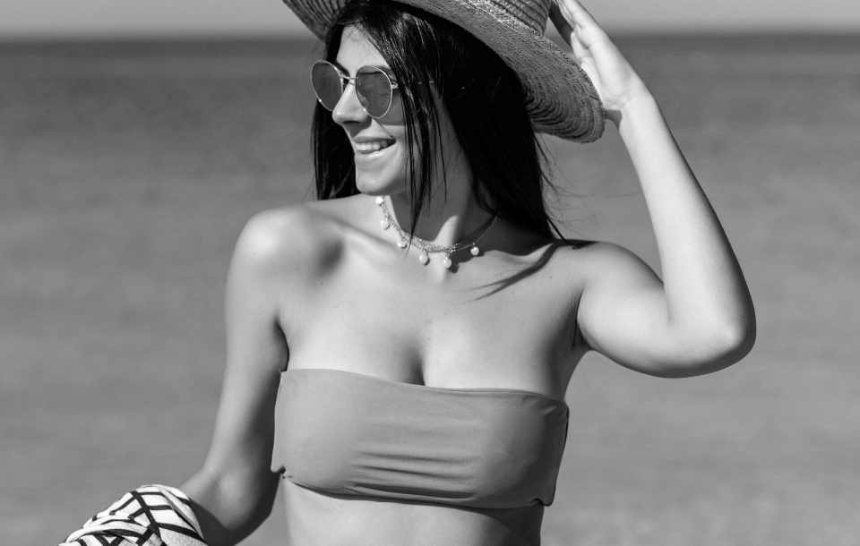 Woman wearing a strapless bikini top and wide-brimmed hat, smiling while holding the brim.