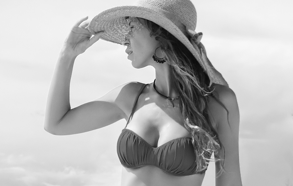 Woman in a bikini and wide-brimmed hat looking to the side with the ocean behind her.