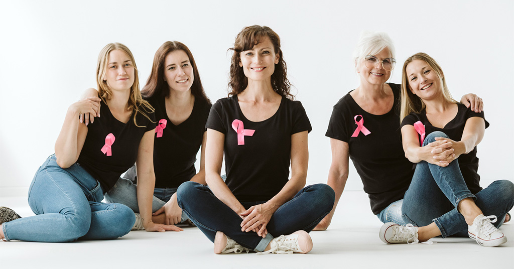 Group of women of different ages sitting together wearing black shirts with pink ribbons.
