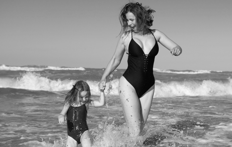 Mother and young daughter playing together in the ocean waves, holding hands and smiling.