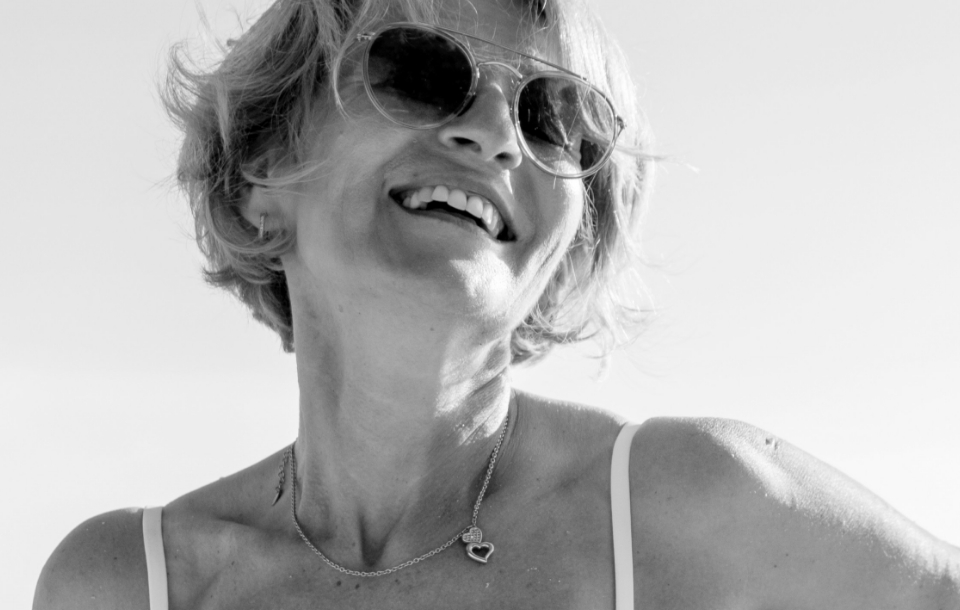 Woman with short hair and sunglasses smiling joyfully in bright sunlight.