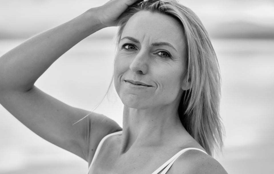 Woman standing by the water with one hand in her hair, giving a calm, confident expression.