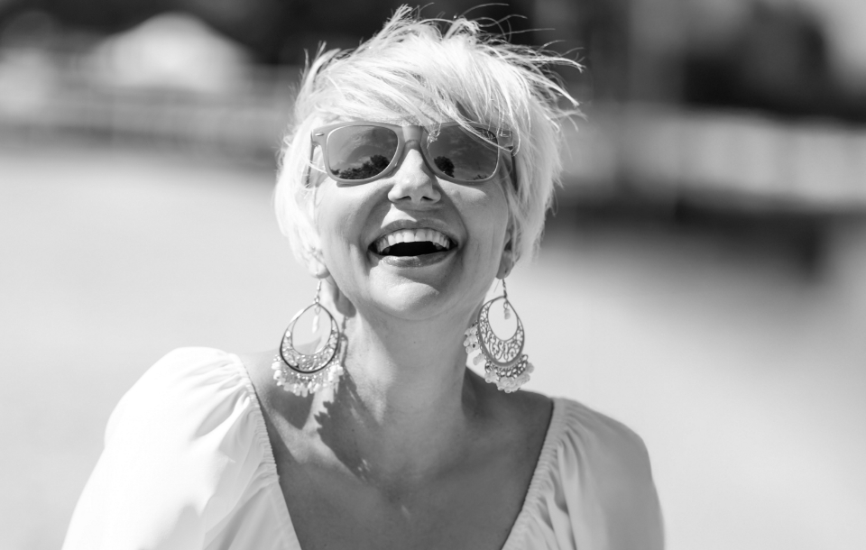 Woman with short blonde hair and large earrings laughing in the sunshine at the beach.