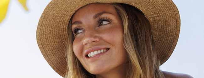 Woman wearing a wide sun hat smiling upward, with luminous, refreshed skin.