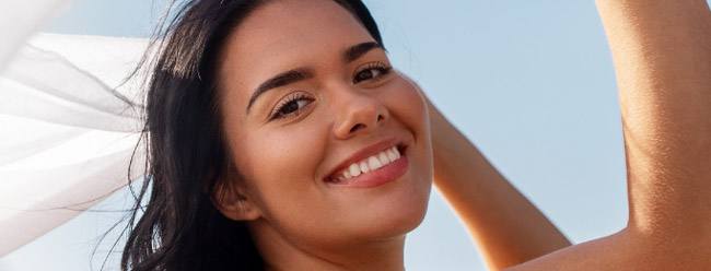 Woman smiling brightly and holding a white scarf against a clear sky.