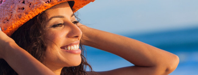 Woman smiling widely and holding a woven orange hat with the ocean behind her.