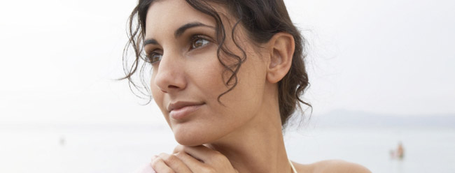 Woman with dark curly hair gazing thoughtfully to the side near calm ocean water.