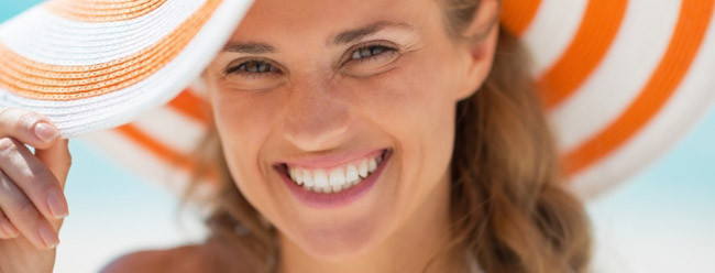 Woman smiling brightly under an orange-and-white sunhat on a sunny beach.