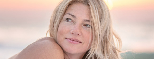 Blonde woman relaxing on a beach at sunset with a gentle smile.