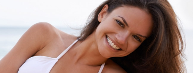 Woman with long dark hair smiling brightly while leaning on the sand by the ocean.