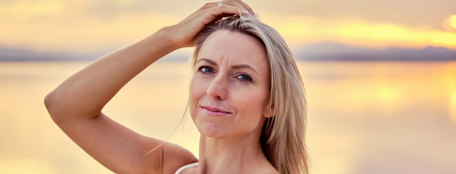 Woman with blonde hair touching her head while standing by the water at sunset.