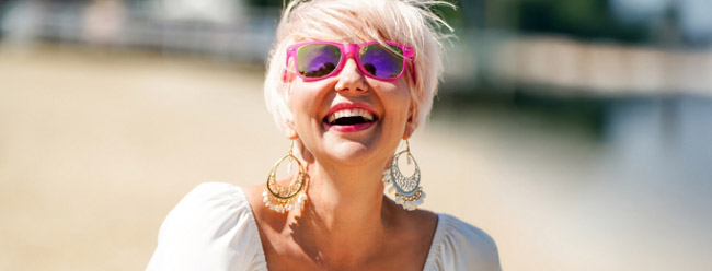 Woman with short blonde hair and pink sunglasses laughing brightly on a sunny beach.