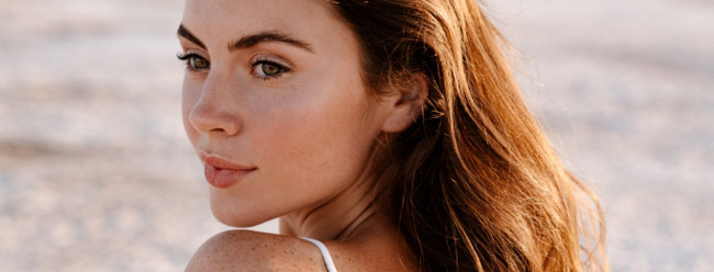 Young woman with light brown hair looking over her shoulder on a sunlit beach.