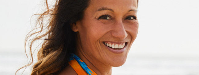 Smiling woman with windblown hair standing by the beach in a colorful top.