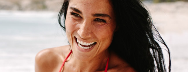 Woman laughing warmly with long dark hair and a red swimsuit by the shore.