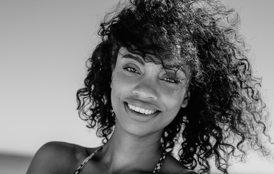 Woman with curly hair smiling in the sun, highlighting her bright, healthy complexion.