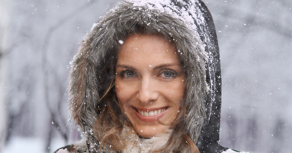 Smiling woman wearing a fur-lined hood outdoors while snow falls around her.