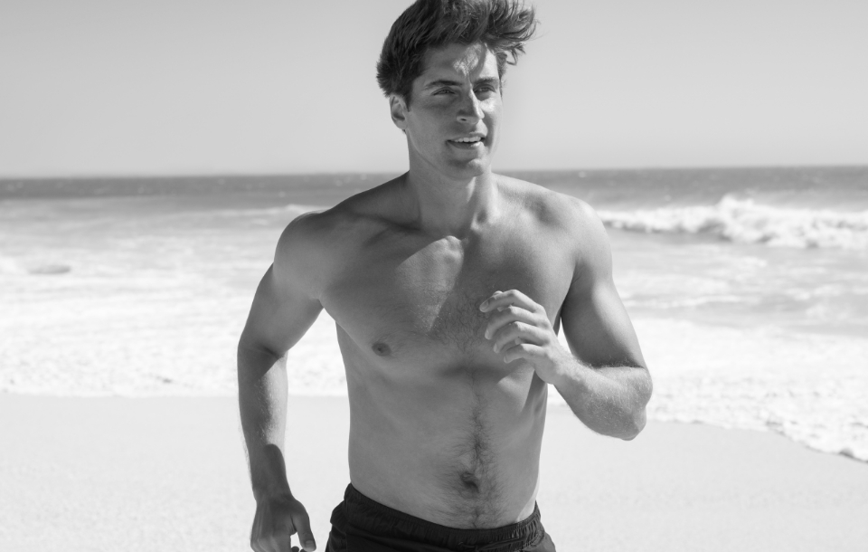 Shirtless man jogging along the shoreline with ocean waves in the background.
