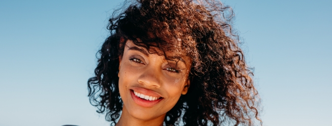 Woman with curly hair smiling in the sun, highlighting her bright, healthy complexion.
